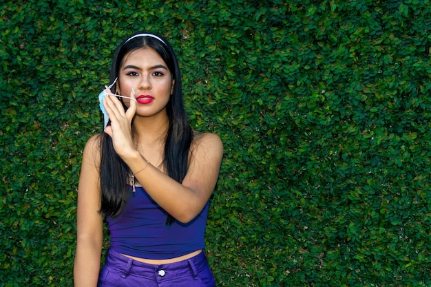 Outdoor portrait of young latin woman removing a mask in front of a leaf wall