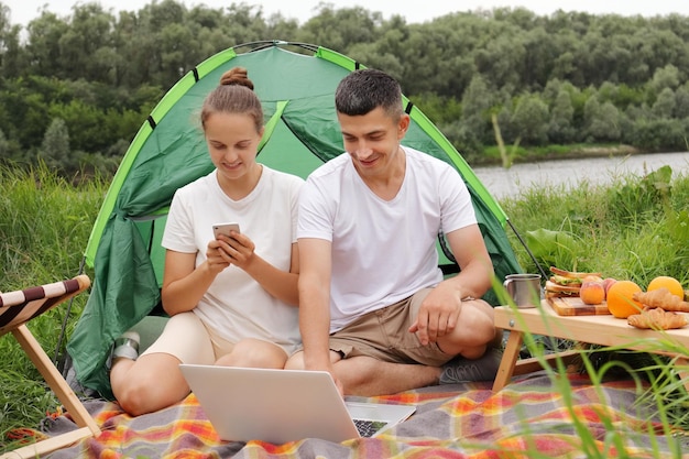 Outdoor portrait of young pretty hiker couple sitting near a tent looking at laptop in the nature woman using cell phone family spending time in open air enjoying resting