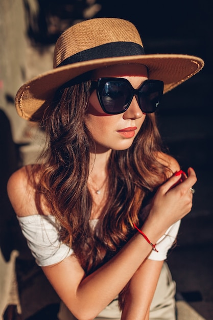 Outdoor Portret van een jonge vrouw die vintage outfit en accessoires draagt. Meisje met bril en hoed zittend op de trap bij zonsondergang