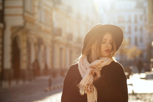 Outdoor portret van fantastische blonde vrouw met hoed, sjaal en jas, poseren in de oude straat in schittering van de zon. Ruimte voor tekst