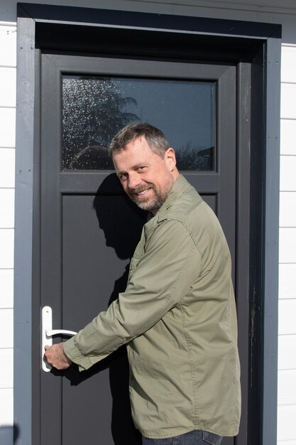 Outdoor portret van gelukkige knappe blanke man open grijze deur houten hut achtergrond huis