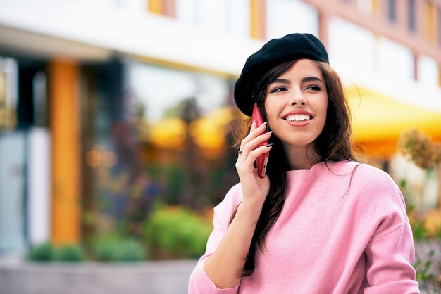 Outdoor Portret van gelukkige vrouw glimlachend en staande buitenshuis praten op mobiele telefoon Jonge brunette vrouw draagt roze trui zwarte baret rusten buiten in de stad wating haar vrienden in de stad