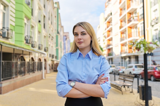 Outdoor Portret van jonge zelfverzekerde zakenvrouw met gekruiste armen, stad straat achtergrond. Vrouwelijke kantoormedewerker in blauw shirt kijkt naar de camera