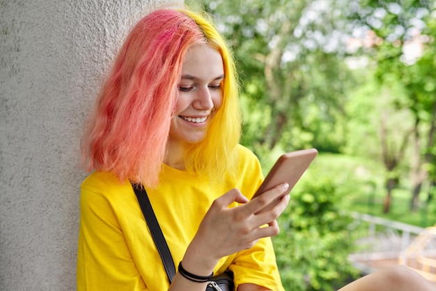 Outdoor Portret van tiener hipster meisje met geverfd gekleurd haar met smartphone in haar handen glimlachend modieuze heldere jonge vrouw in gele tshirt met geel roze haren