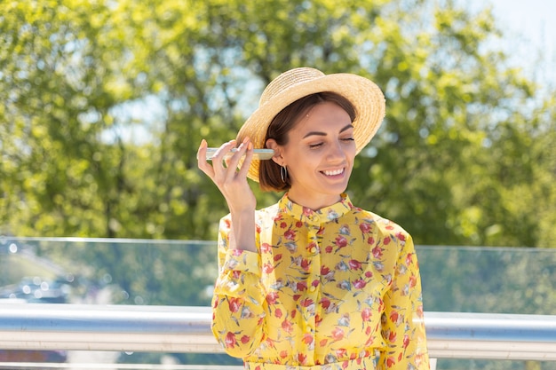 Outdoor portret van vrouw in gele zomerjurk en hoed luisteren audio spraakbericht op telefoon