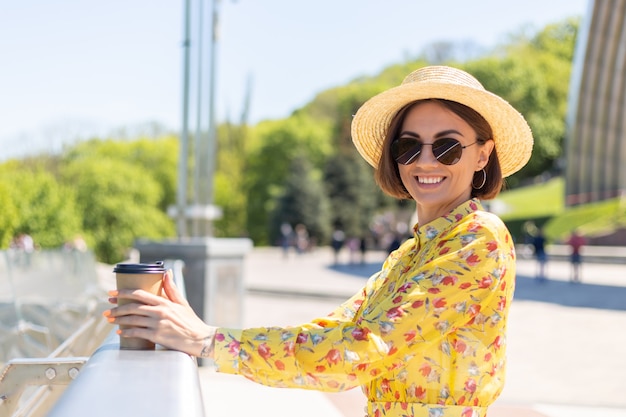 Outdoor Portret van vrouw in gele zomerjurk en hoed met kopje koffie genieten van zon, staat op brug met prachtig uitzicht op de stad