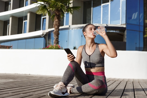 Открытый выстрел привлекательной фитнес-женщины, сидящей на деревянном пирсе со смартфоном