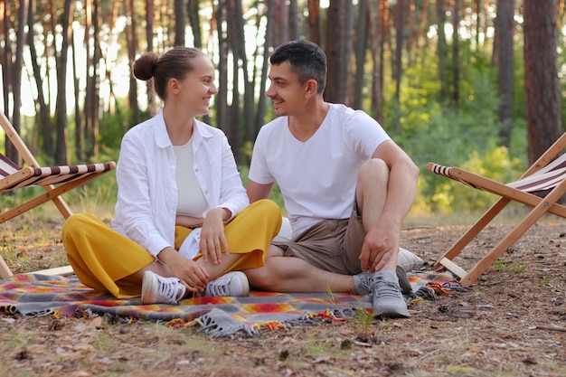 Photo outdoor shot of beautiful young couple sitting on a plaid in an summer park looking at each other with love and gentle expressing positive emotions having vacation in the forest