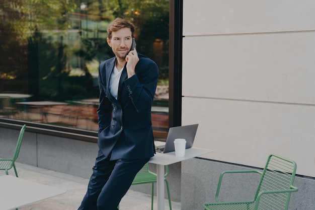 Outdoor shot of busy man entrepreneur solves probems distantly has telephone conversation poses in open air cafe near table with laptop computer and coffee wears formal suit discusses something