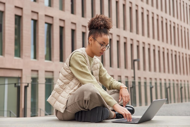 Photo outdoor shot of clever female student does remote job via laptop computer sits crossed legs wears casual clothes poses against modern building woman blogger chatswith friends in social networks