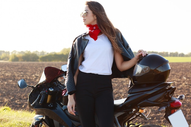 Outdoor shot of confident risky young woman standing near her motorbike