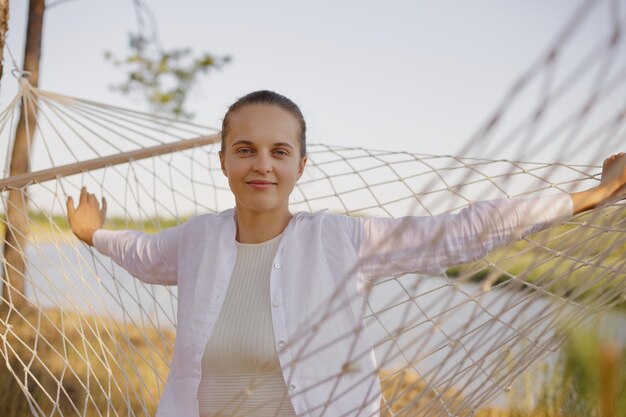 Outdoor shot of dark haired Caucasian woman wearing white shirt sitting in hammock and looking at camera spending her free time outdoor near water posing on the bank of the river