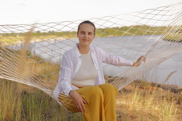 Outdoor shot of good looking pretty female wearing white shirt sitting in comfortable hammock on the bank of the river looking at camera resting by the water