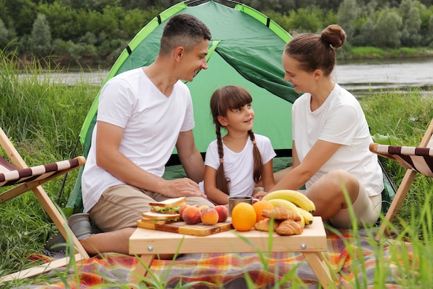 Colpo all'aperto di una famiglia felice seduta vicino alla tenda a terra che fa un picnic insieme godendosi frutta deliziosa e panini parlando e trascorrendo un felice fine settimana