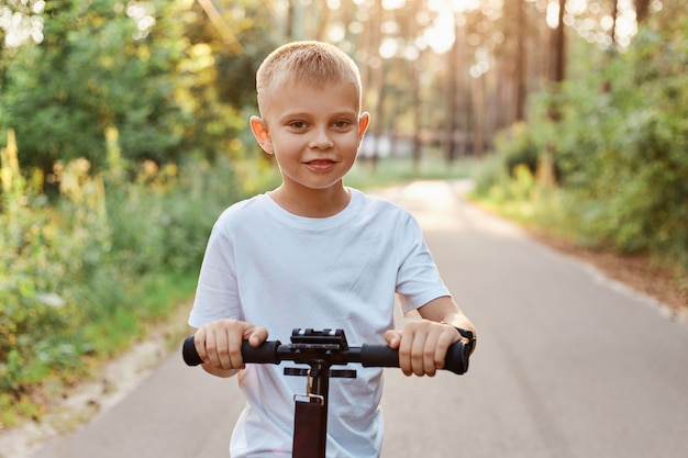 Photo outdoor shot of happy little blond boy playing with his scooter at asphalt road in summer park, cute kid wearing white casual t shirt, happy childhood, healthy lifestyle.