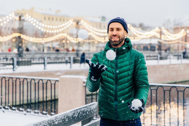 Outdoor shot of happy smiling bearded male in warm jacket and hat plays snowballs as stands against beautiful lights being in good mood has joyful expression People winter and fun concept