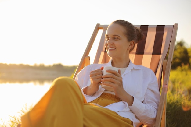 Outdoor shot of happy smiling dark haired female wearing white t shirt sitting in folding chair near the river looking away with delighted expression enjoying coffee or tea