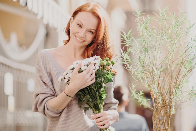 Outdoor shot of lovely foxy woman tilts head happy to recieve flowers from boyfriend dressed in casual clothes has date wth boyfriend Attractive red haired female likes spring time Street style