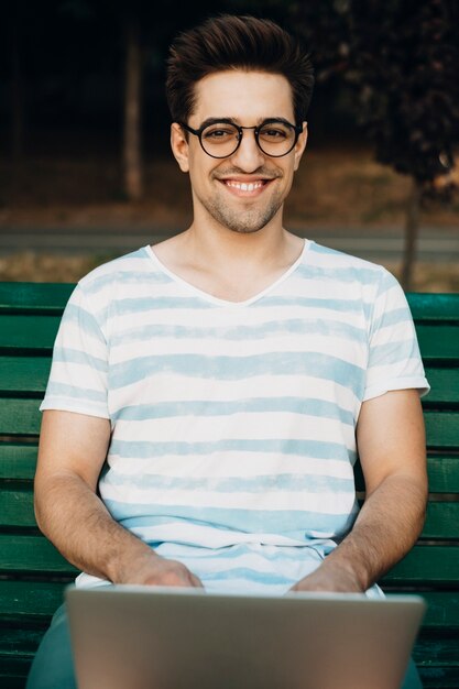 Photo outdoor shot of a man vlogger looking at camera smiling. male influencer sitting on a bench with a laptop on his legs.