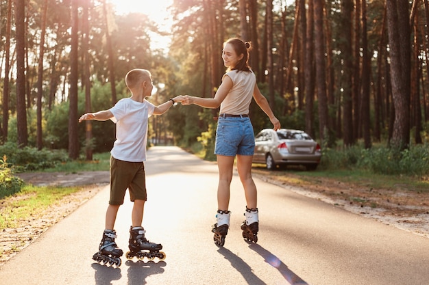 Outdoor shot of mother and son on roller skates having together, woman with her child dancing while rollerblading in summer park, happy family spending tome in nature in actively.