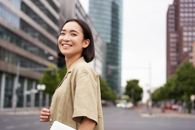 写真 街の中心部のどこかに行くラップトップを持つアジアの女の子の屋外ショットは、街に行く通りを歩いています