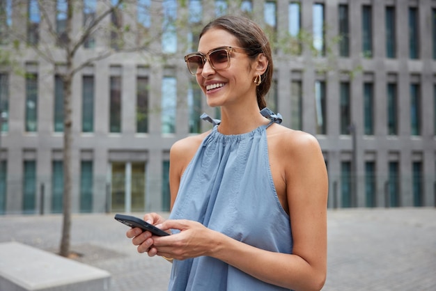 スタイリッシュな女性の屋外ショットはサングラスをかけています青いドレスは都会の設定で歩きますhols携帯電話は友人とのメッセージを楽しんでいます