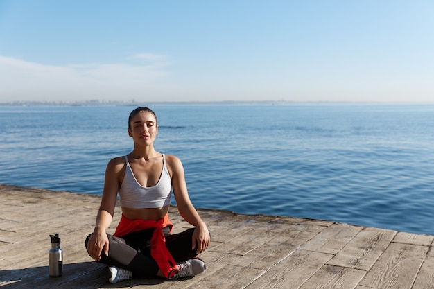 Outdoor shot of peaceful fitness woman sitting in asana and doing yoga near the sea.