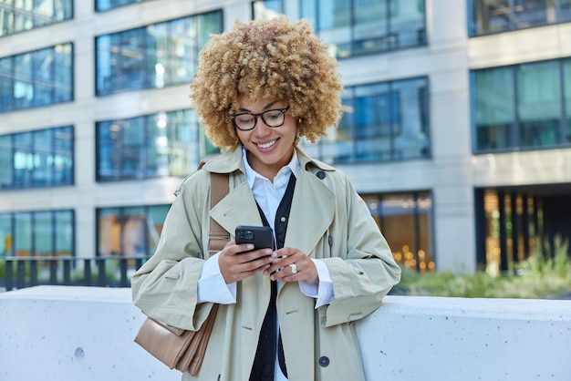 Outdoor shot of pretty beautiful curly haired young woman uses\
smartphone technology for writing web review browses website wears\
spectacles and raincoat carries bag poses against urban\
building