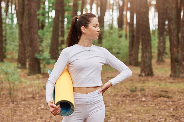 Outdoor shot of slim confident female in white sportswear, holding yoga mat in hands, looking away and keeping hands on hips