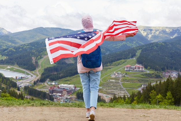 Outdoor shot of slim girl at nature with American flag on her shoulders, woman wearing casual rosy jacket, trousers