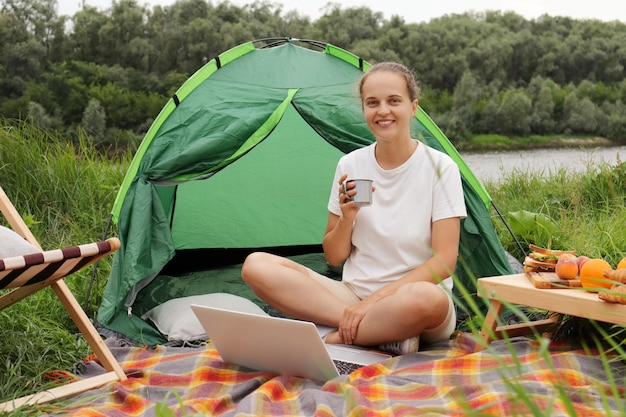 Outdoor shot of smiling happy woman freelancer sitting on the ground on blanket and drinking coffee or tea working online while having rest in nature near river