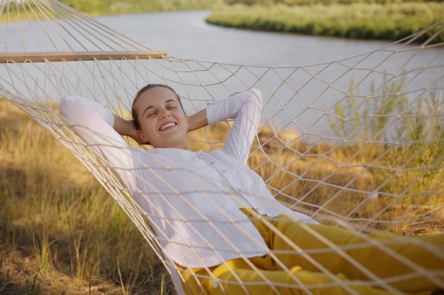 Outdoor shot of smiling relaxed young adult woman sitting on hammock by the water with raised arms looking at camera with toothy smile enjoying resting in nature