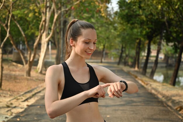 Outdoor shot of sporty caucasian woman tracking fitness results on smartwatch poses outdoors