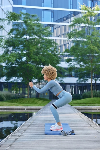 Outdoor shot of sporty woman dressed in blue tracksuit does\
squats with resistance band has workout near lake poses against\
urban background people healthy lifestyle and exercising\
concept