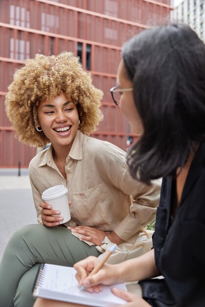 Outdoor shot of two happy diverse female students meet together\
discuss future project write down information in notebook drink\
coffee to go laugh joyfully have good mood cooperation concept