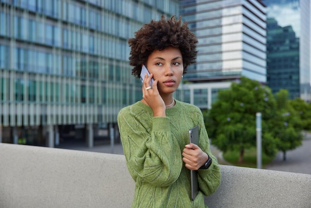 Outdoor shot van serieuze jonge vrouw belt houdt tablet terloops gekleed brengt vrije tijd door in stedelijke omgeving maakt gebruik van moderne technologieën poses in stedelijke omgeving geniet van mobiliteitscommunicatie