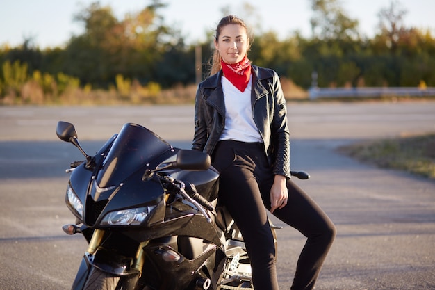 Outdoor shot of woman with dark hair sitting on black motorbike