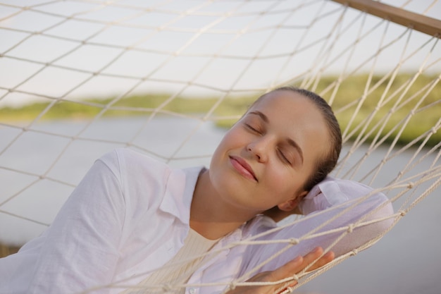 Outdoor shot of young adult caucasian female wearing white\
shirt lying on hammock on the bank of the river lying with closed\
eyes resting and sleeping by the water