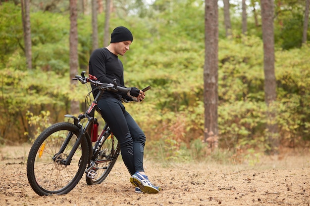 Outdoor shot of young European rider standing in forest surrounded with trees, holding mobile phone