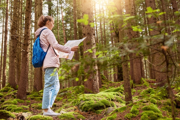 Outdoor shot of young slender backpacker being inspired by travelling, enjoying active rest, holding map, following trip way, good at orientating, wearing casual clothes. Leisure activities concept.