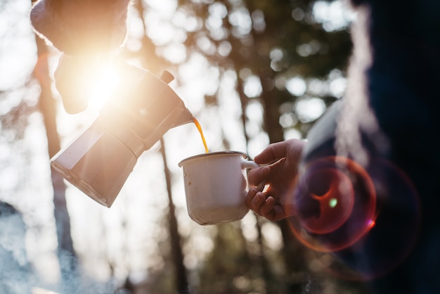 Открытый выстрел молодой женщины наливает себе горячий напиток в горах рядом с костром во время заката.