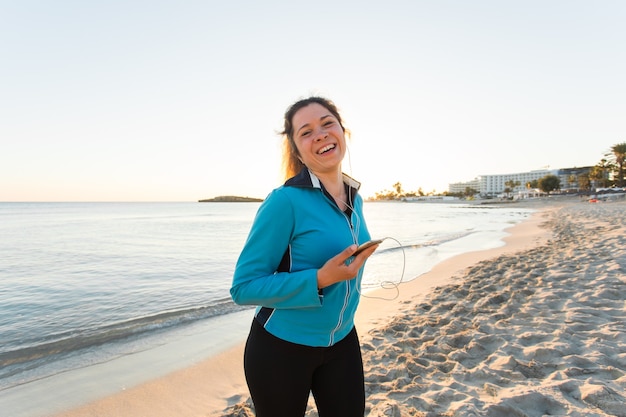 Sport all'aperto, gadget per il fitness e concetto di persone - fitness femminile sorridente che tiene in mano uno smartphone con gli auricolari.