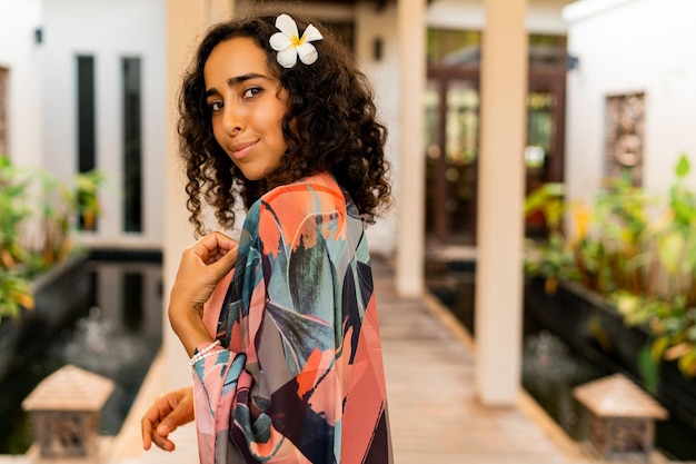 Outdoor summer portrait of pretty brunette woman with tropical flower in hairs posing in  stylish  hotel