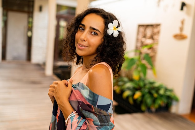 Foto ritratto estivo all'aperto di bella donna bruna con fiore tropicale nei capelli in posa in un hotel elegante