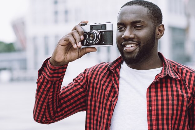 Outdoor summer smiling lifestyle portrait of handsome and happy Afro American tourist having fun in the city in Europe with camera travel photo of photographer making pictures in hipster style