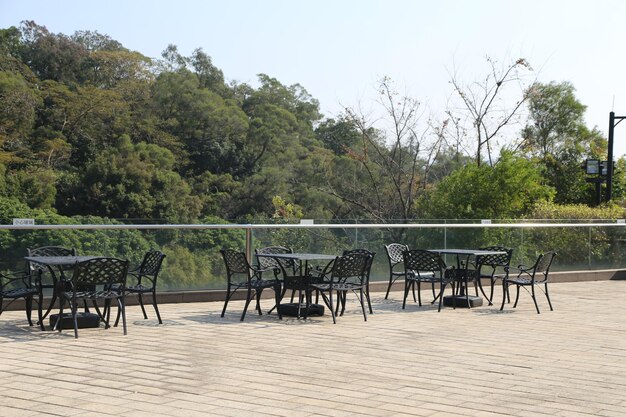 the outdoor terrace of the restaurant has black iron tables and chairs with an overhead glass fence overlooking green trees