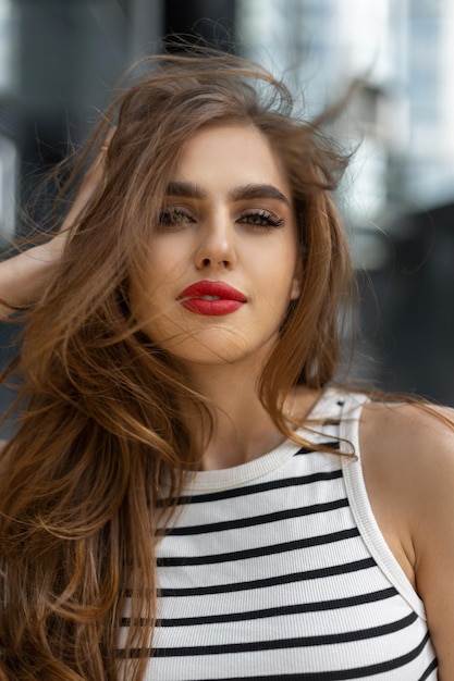 Outdoor urban female portrait of a beautiful stylish fresh woman with red lips in a striped top walks on a windy day and looks at the camera