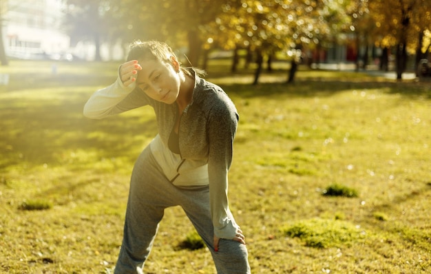 Outdoor workout concept. Tired sport woman in sport clothes wipes sweat from her forehead with her sleeve in the autumn park