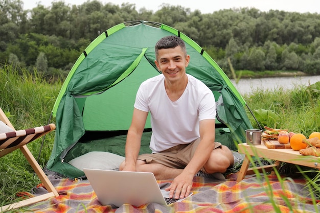 Outdoor young adult man tourist freelancer working online on a\
laptop outdoors sitting on ground near tent looking at camera with\
happy satisfied expression