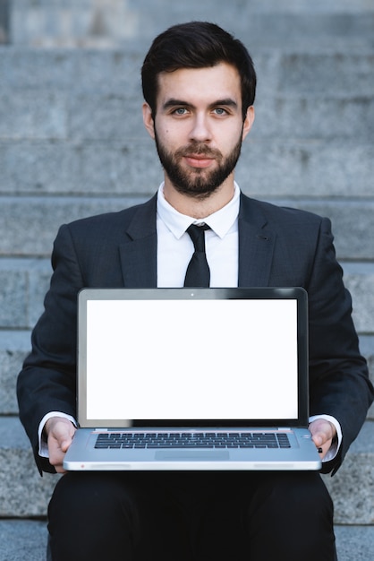 Outdoors businessman sitting on the steps with a laptop on his lap and looking forward. 